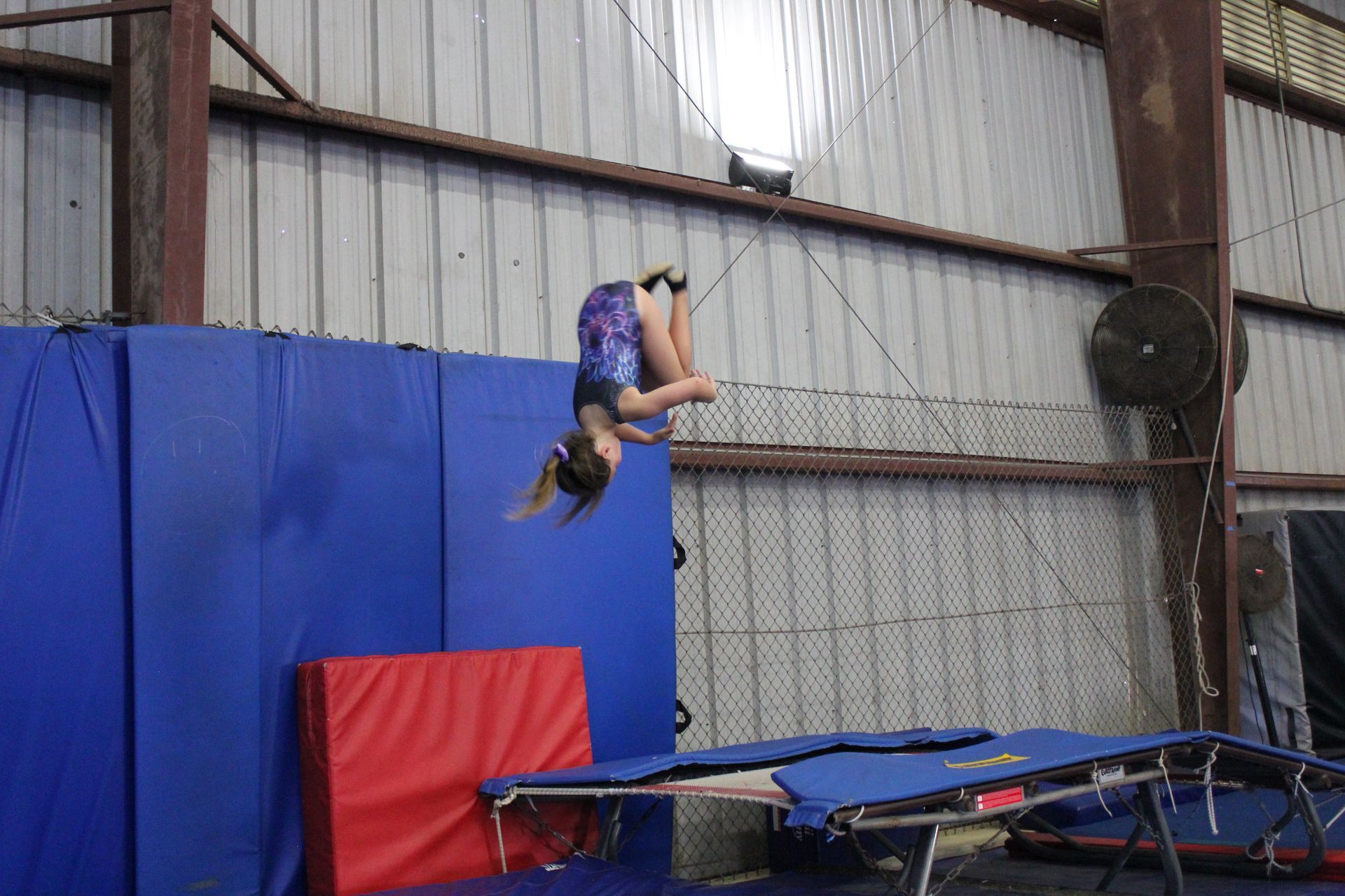 Girl doing a front flip on a trampoline, in a gym with mats and blue padding.