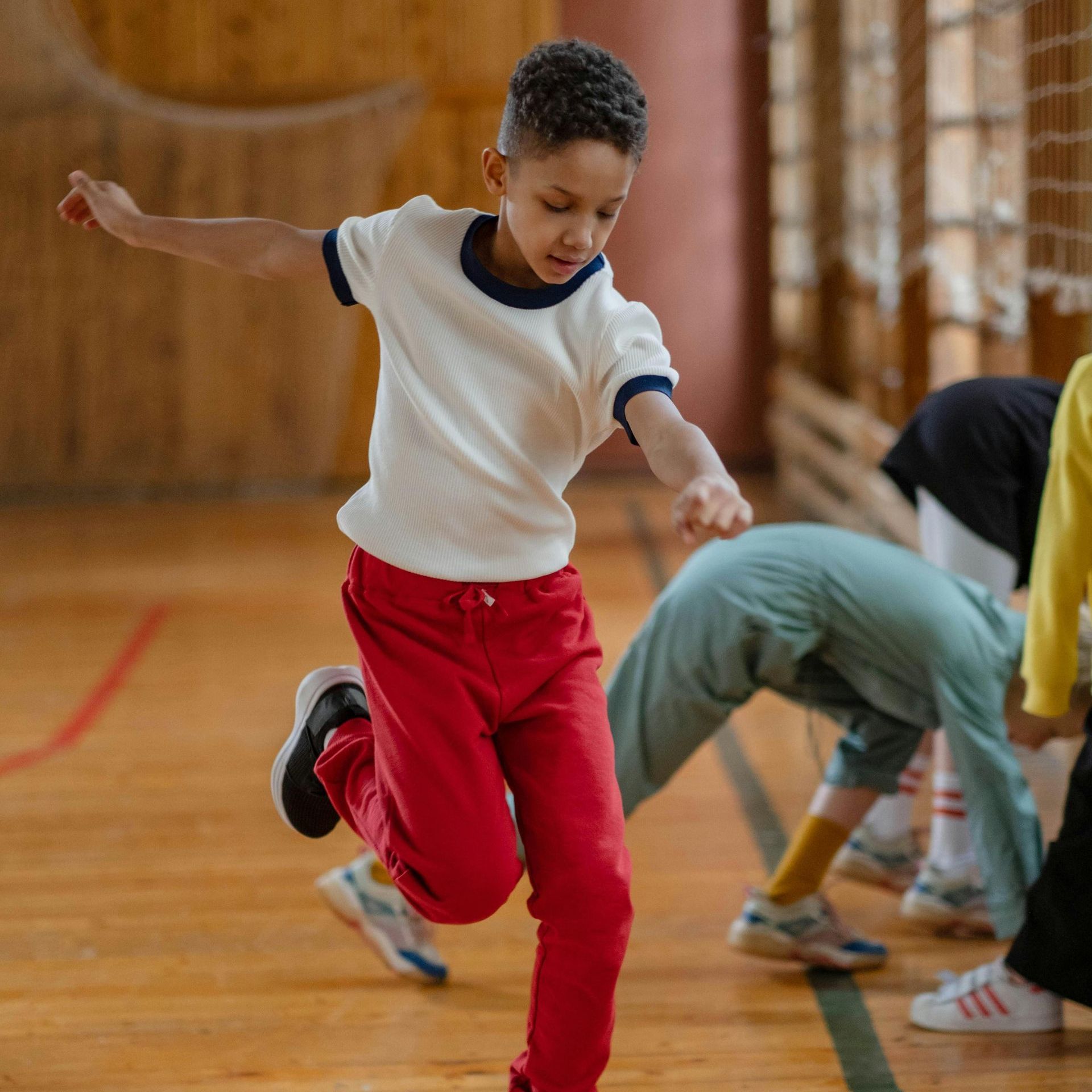 A young boy in a white shirt and red pants is running on a wooden floor.
