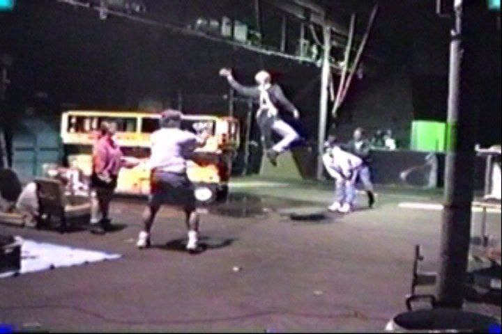 A person in a suit jumps in the air on a stage as others watch in an outdoor, nighttime setting with a bus backdrop.