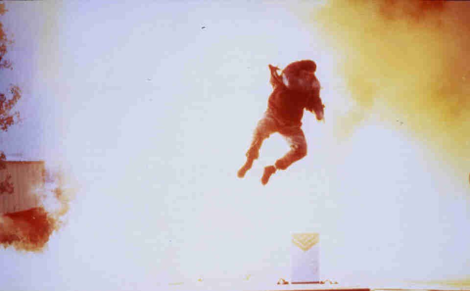 A person in dark clothing is propelled through the air by a bright explosion in a bright, hazy outdoor setting.