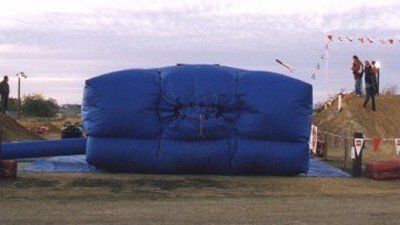 A large, blue rectangular inflatable stunt bag sits on a dirt surface between two ramps, with spectators nearby.