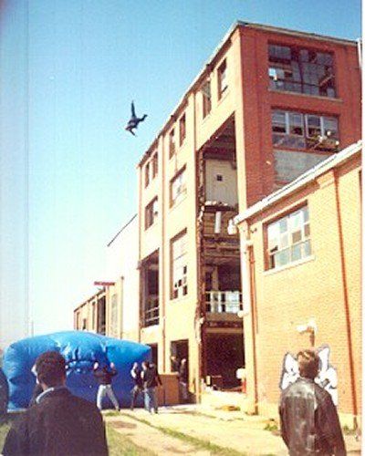 A stunt person jumps from the top floor of a brick building toward a large blue inflatable landing pad below.