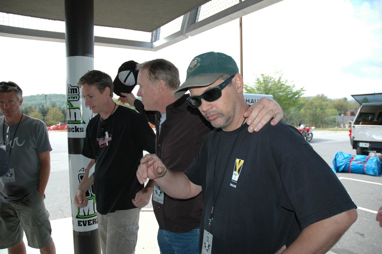 Four people standing under a covered walkway, with one person in a baseball cap and sunglasses putting an arm around another.