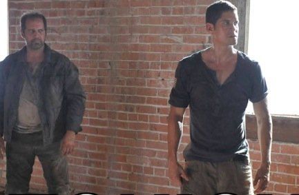 Two men in worn, dusty clothes stand in a room with an exposed brick wall, looking toward the right.
