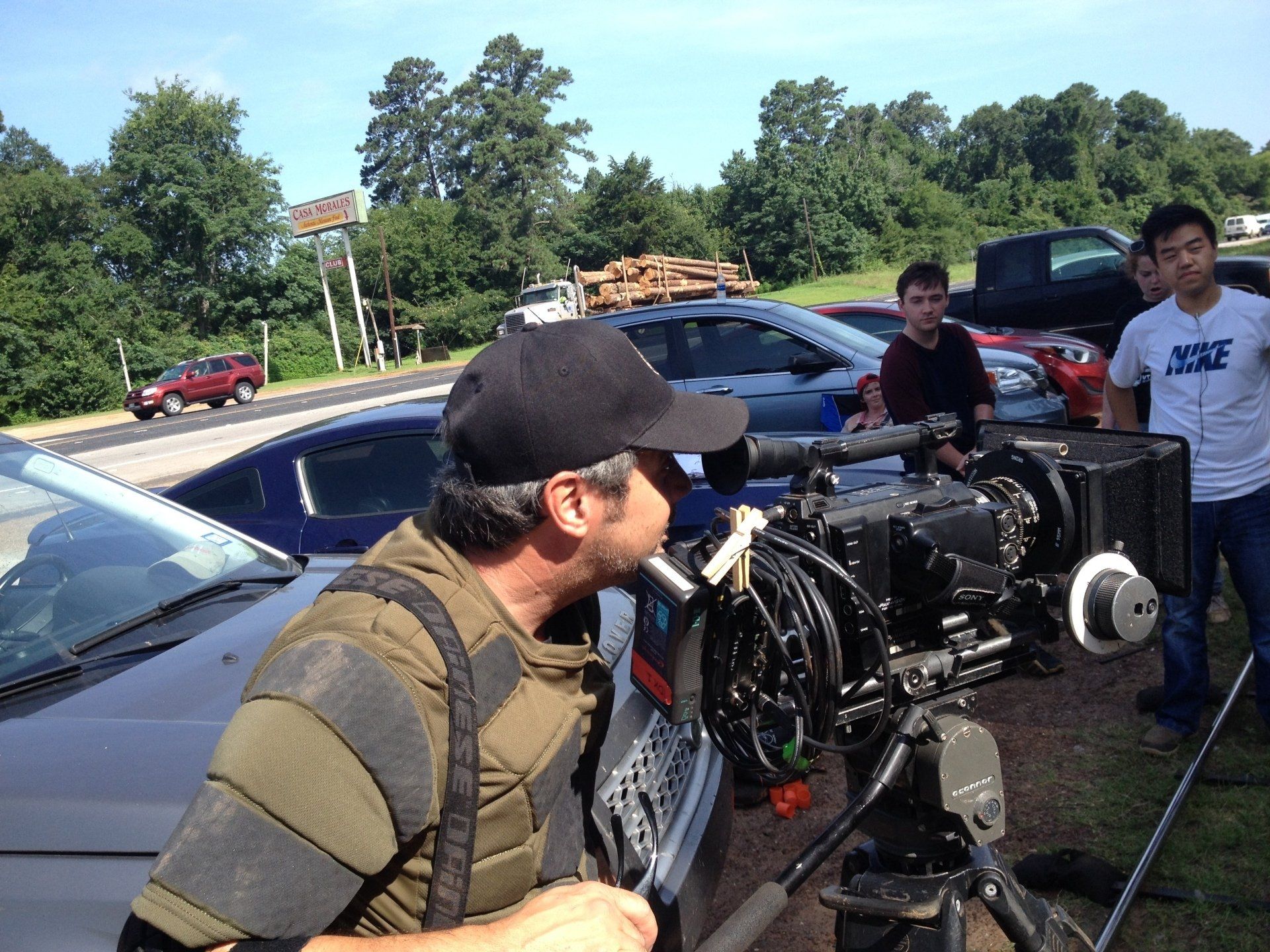 A person in a black cap and tactical vest operates a professional video camera on a tripod outdoors.