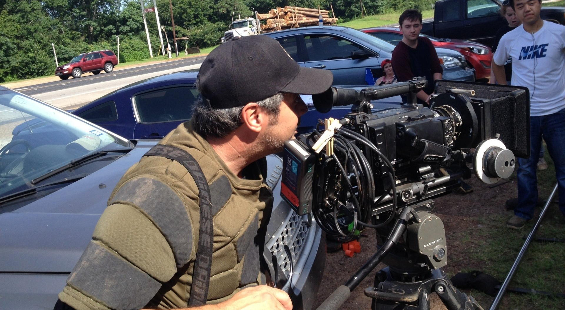 A person in a black cap and tactical vest operates a professional cinema camera mounted on a tripod outdoors.