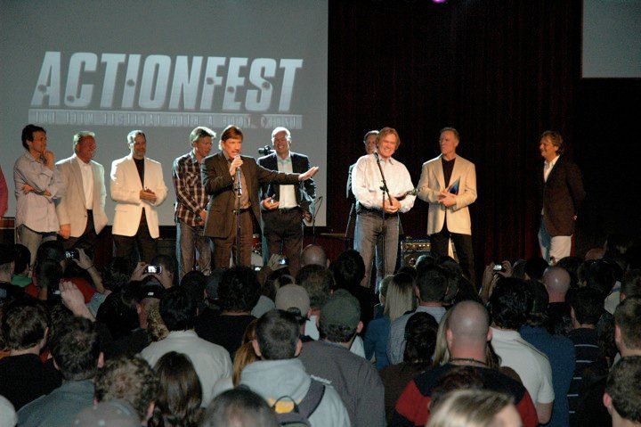 A group of people standing on a stage at an ActionFest event, with one person speaking into a microphone to an audience.