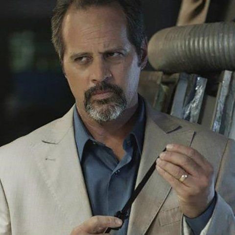 A man with a goatee wearing a beige suit and blue shirt looks down while holding a thin black object.