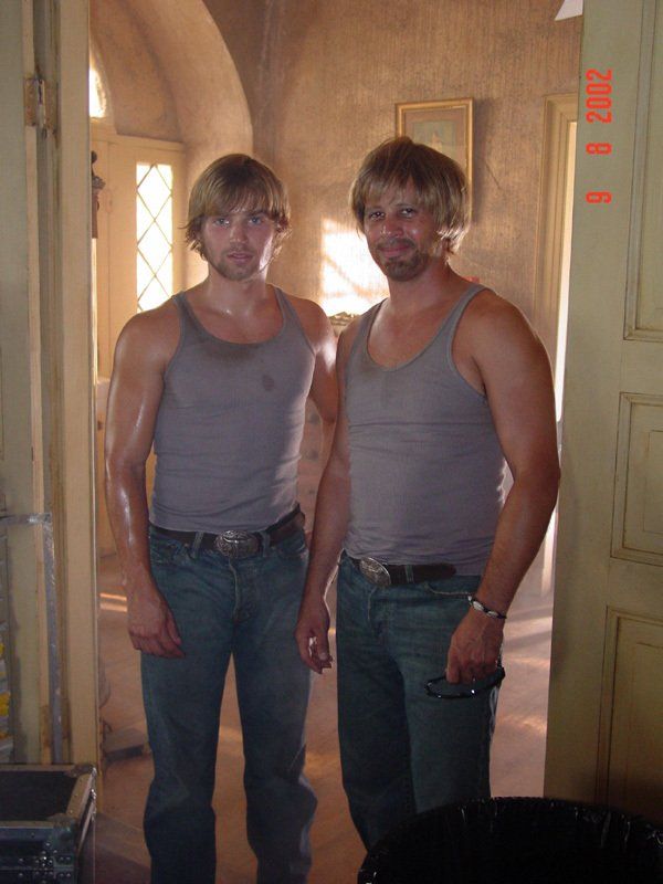 Two people stand side-by-side wearing matching gray tank tops and jeans, posing indoors in a doorway.