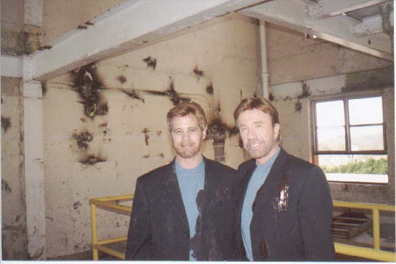 Two men in dark blazers and light blue shirts standing side-by-side in front of a distressed, stained interior wall.