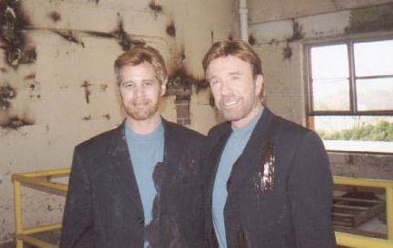 Two men in matching light blue shirts and dark blazers stand smiling side-by-side in a weathered, industrial room.