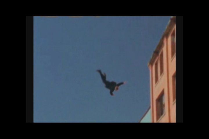 A person in a dark suit falling through the sky alongside the side of a building.