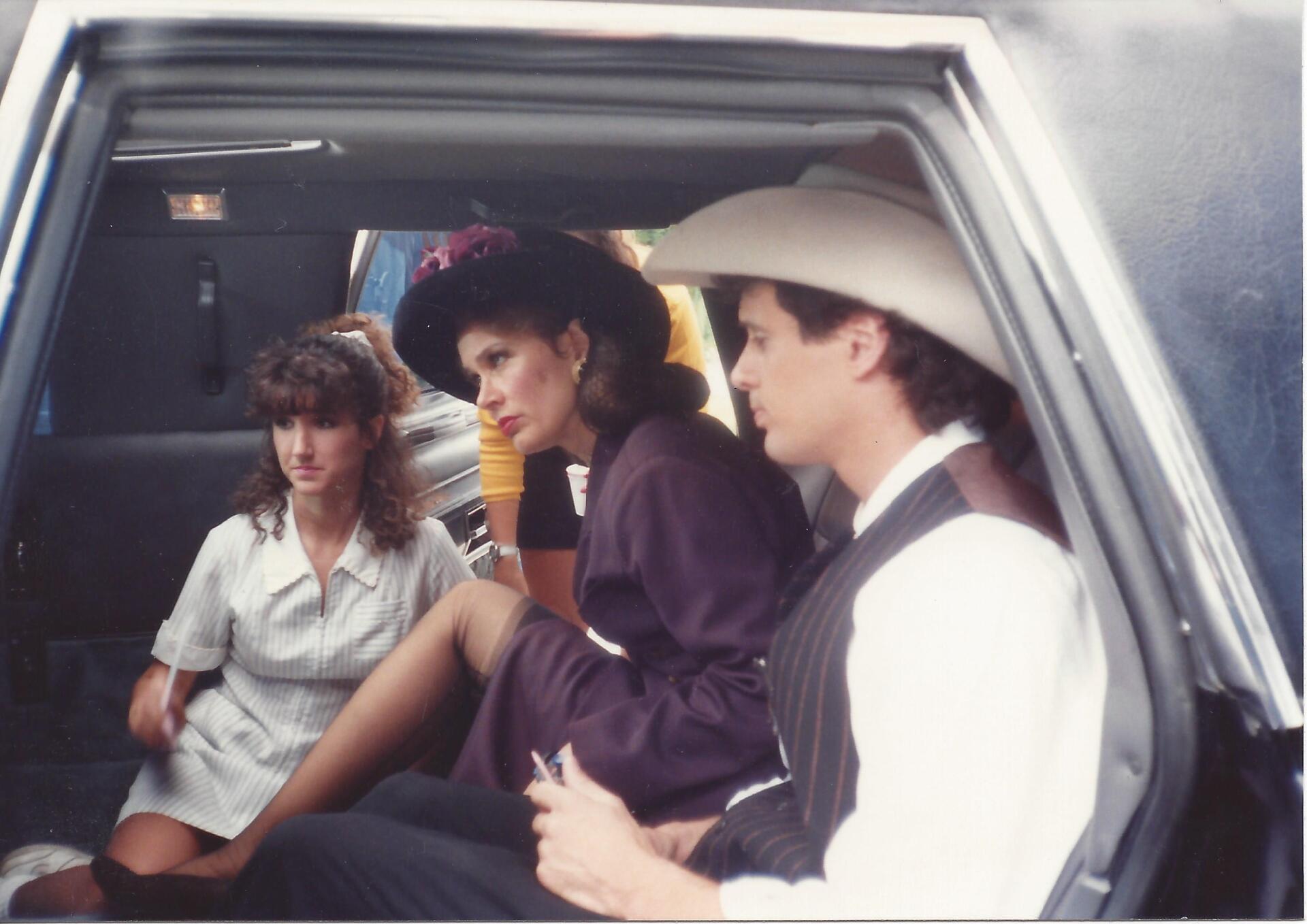 Three people sit in the back seat of a car: a person in a white dress, another in a purple coat and hat, and one in a hat.