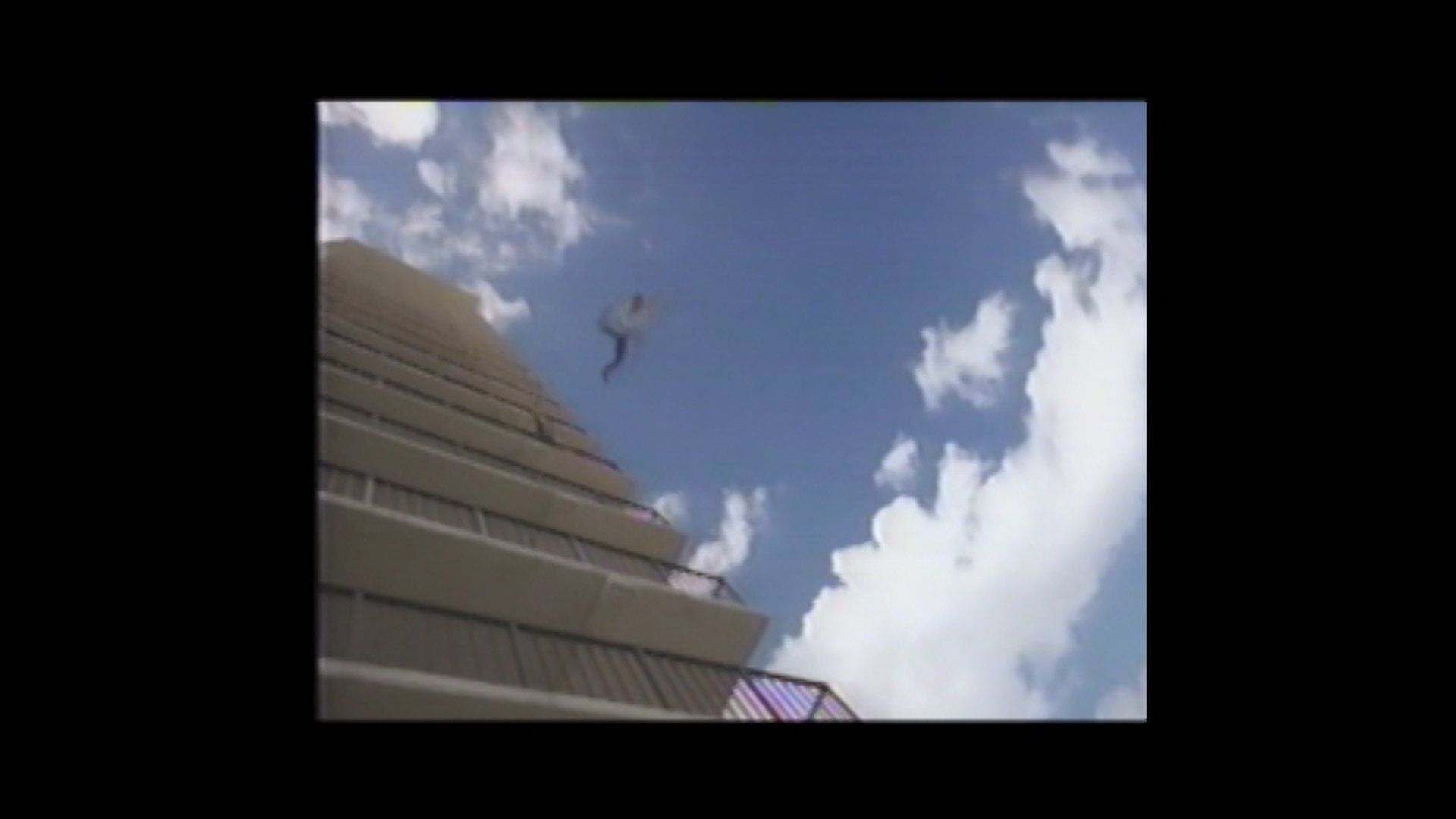 A person jumping from the balcony of a tall concrete building against a bright blue sky with clouds.