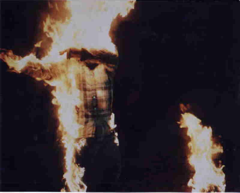 A figure wearing a plaid shirt and dark pants stands engulfed in flames against a dark background.