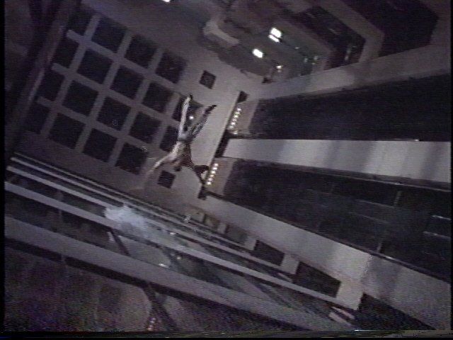 A person falls vertically past the lit windows of a modern building at night, viewed from an angled perspective.