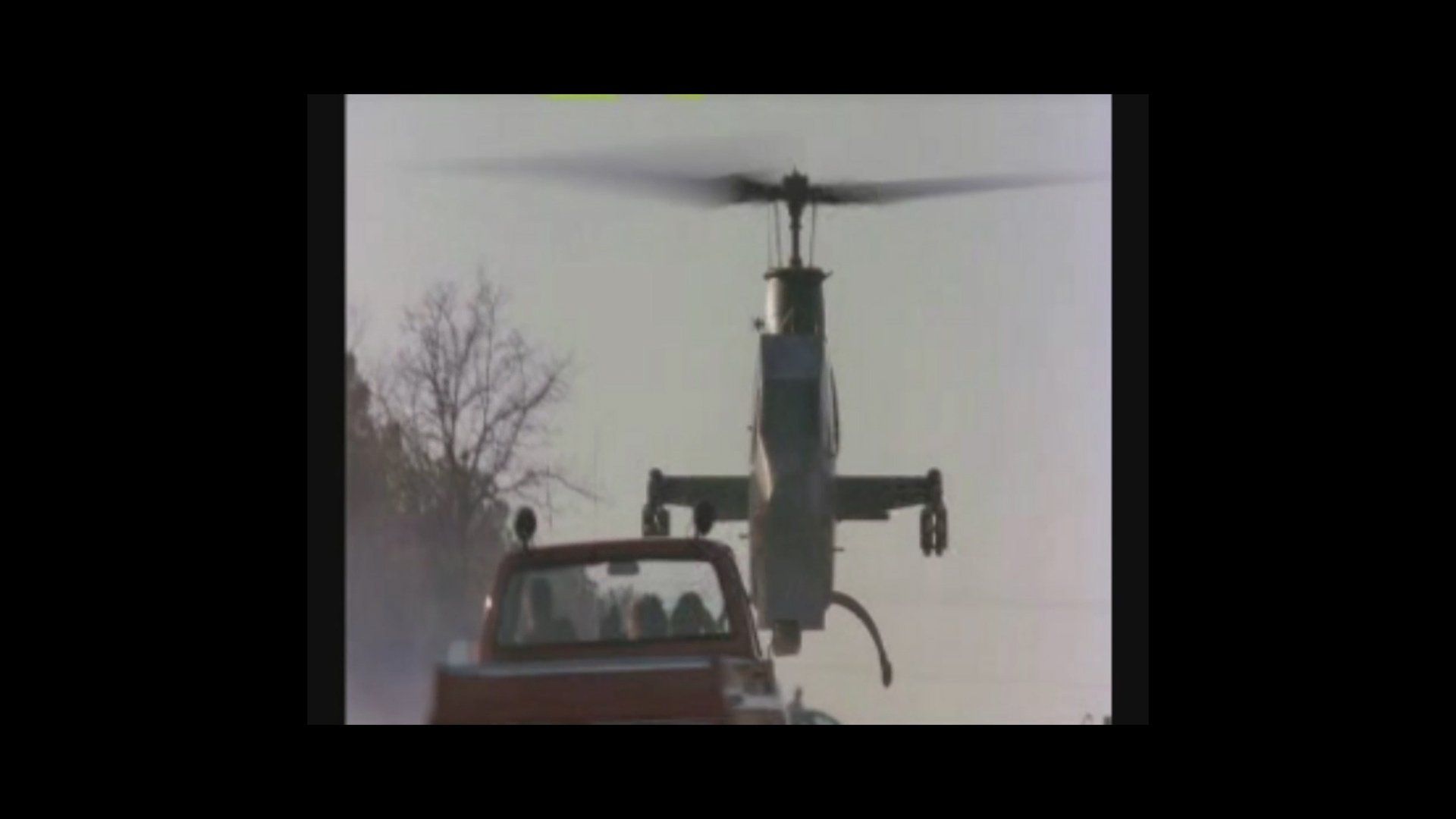 A helicopter flies low behind a red pickup truck on a rural road under a hazy sky.