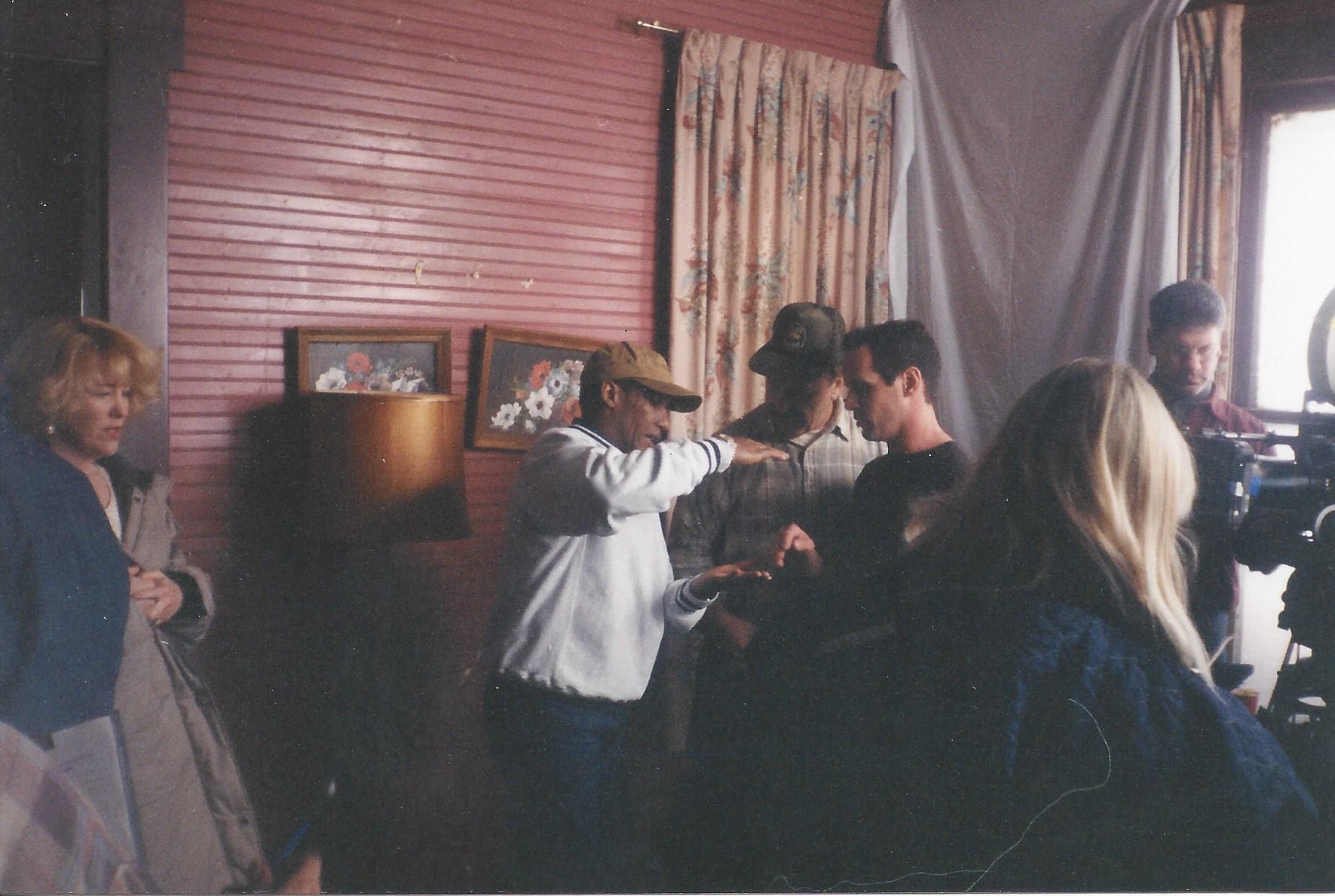 A film crew works in a room with red horizontal paneled walls, featuring a director instructing actors near curtains.