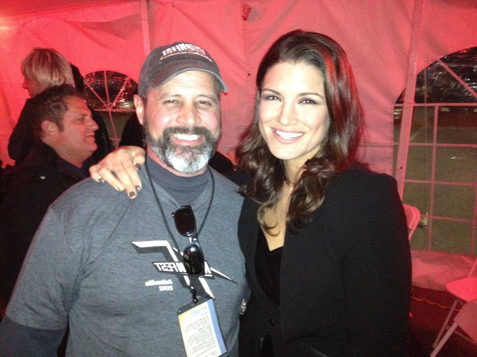 A man wearing a grey t-shirt and baseball cap smiles next to a woman in a black blazer at an evening event.
