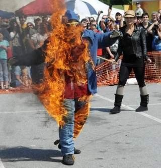 A stunt performer engulfed in flames walks across an outdoor paved area as onlookers watch from behind a fence.