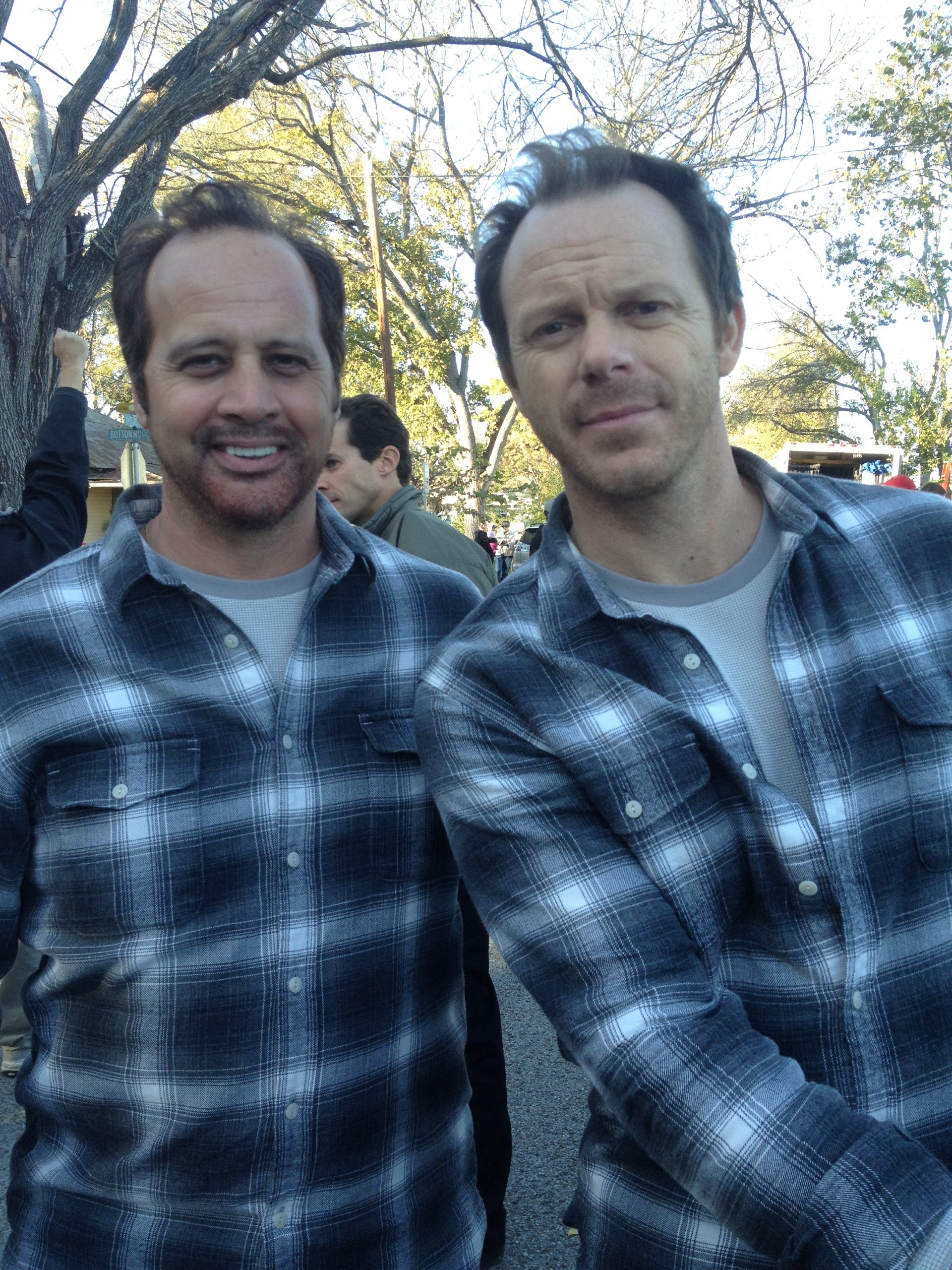 Two men in matching black-and-white plaid flannel shirts pose together outdoors, with trees in the background.