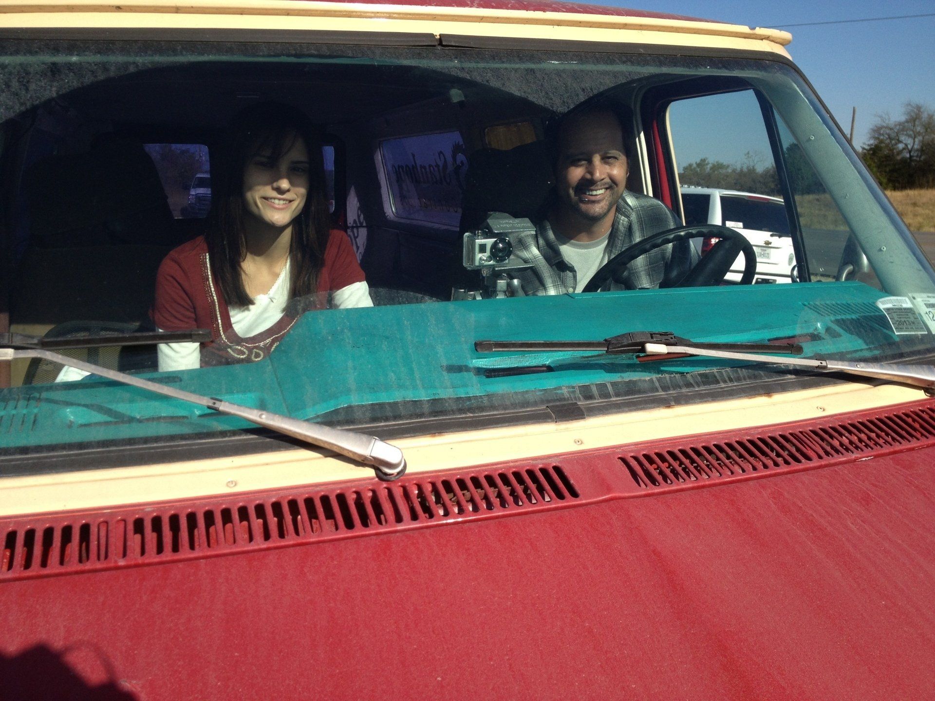 Two people smiling from the front seats of a red van on a sunny day.