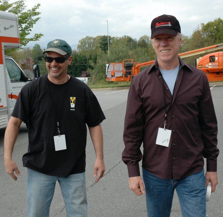 Two men wearing lanyards and hats walk outside near an orange construction lift and a white vehicle.