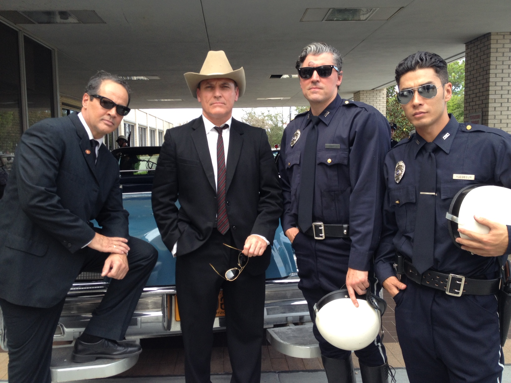 Four people posing by a vintage car; two men in suits and two in police uniforms, all wearing sunglasses.