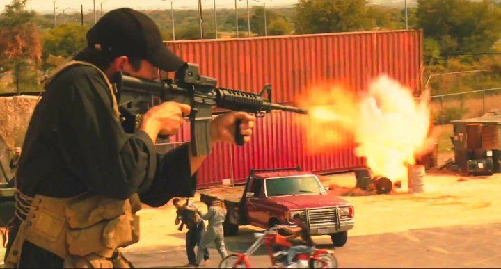 A person in a cap firing an assault rifle toward a large orange shipping container, causing an explosion near a truck.