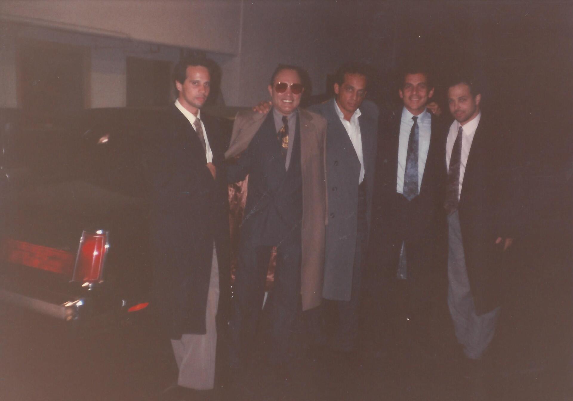 Five people in suits and coats stand in a line together outside at night, next to the rear of a dark vehicle.