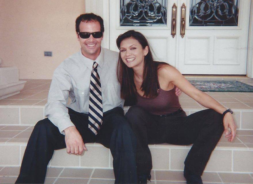 A man in a shirt and tie with sunglasses and a woman in a tank top sit side-by-side on steps outside a building.