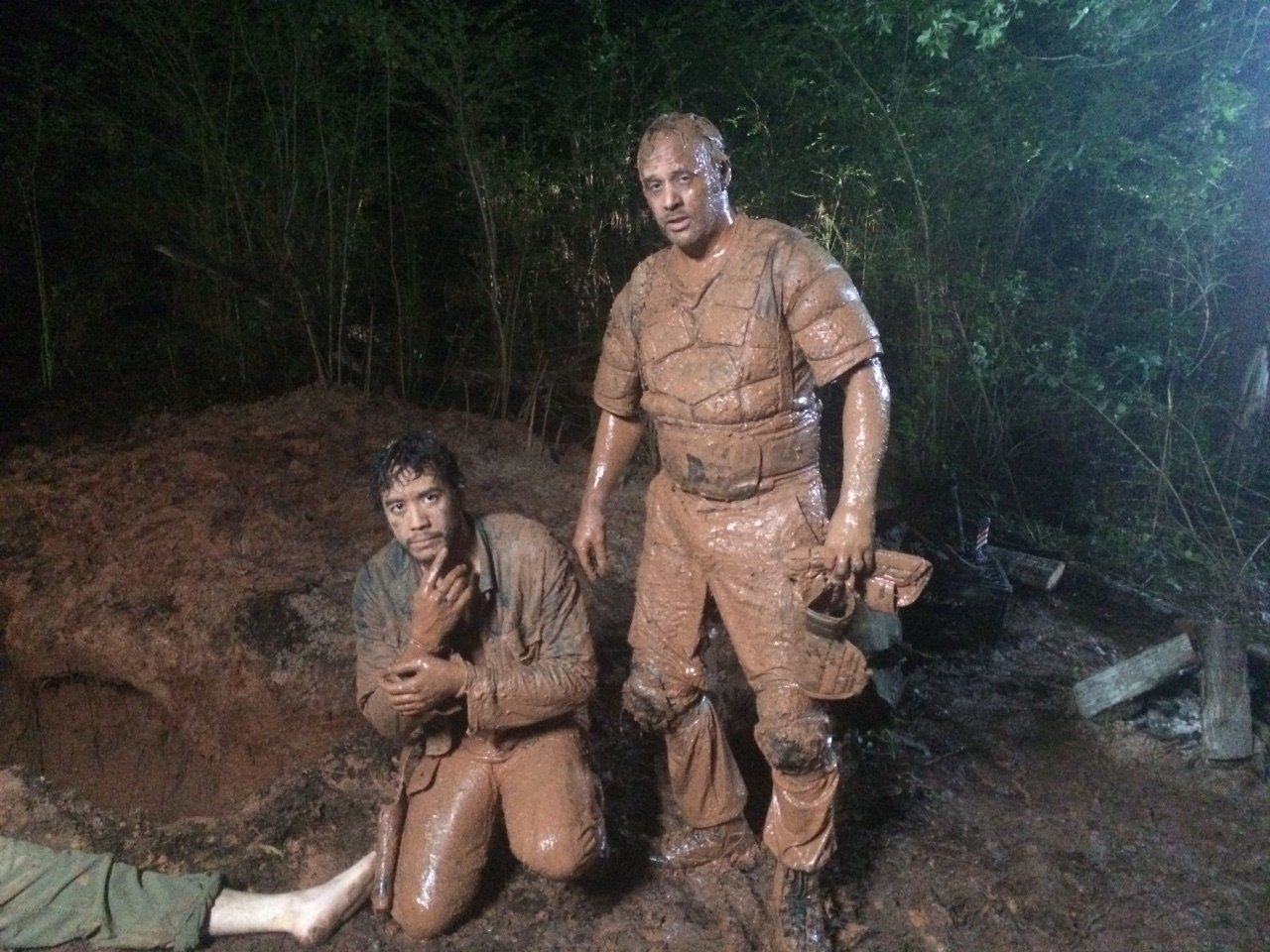 Two people covered in mud stand in a dark, forested area; one is kneeling while the other stands holding a helmet.