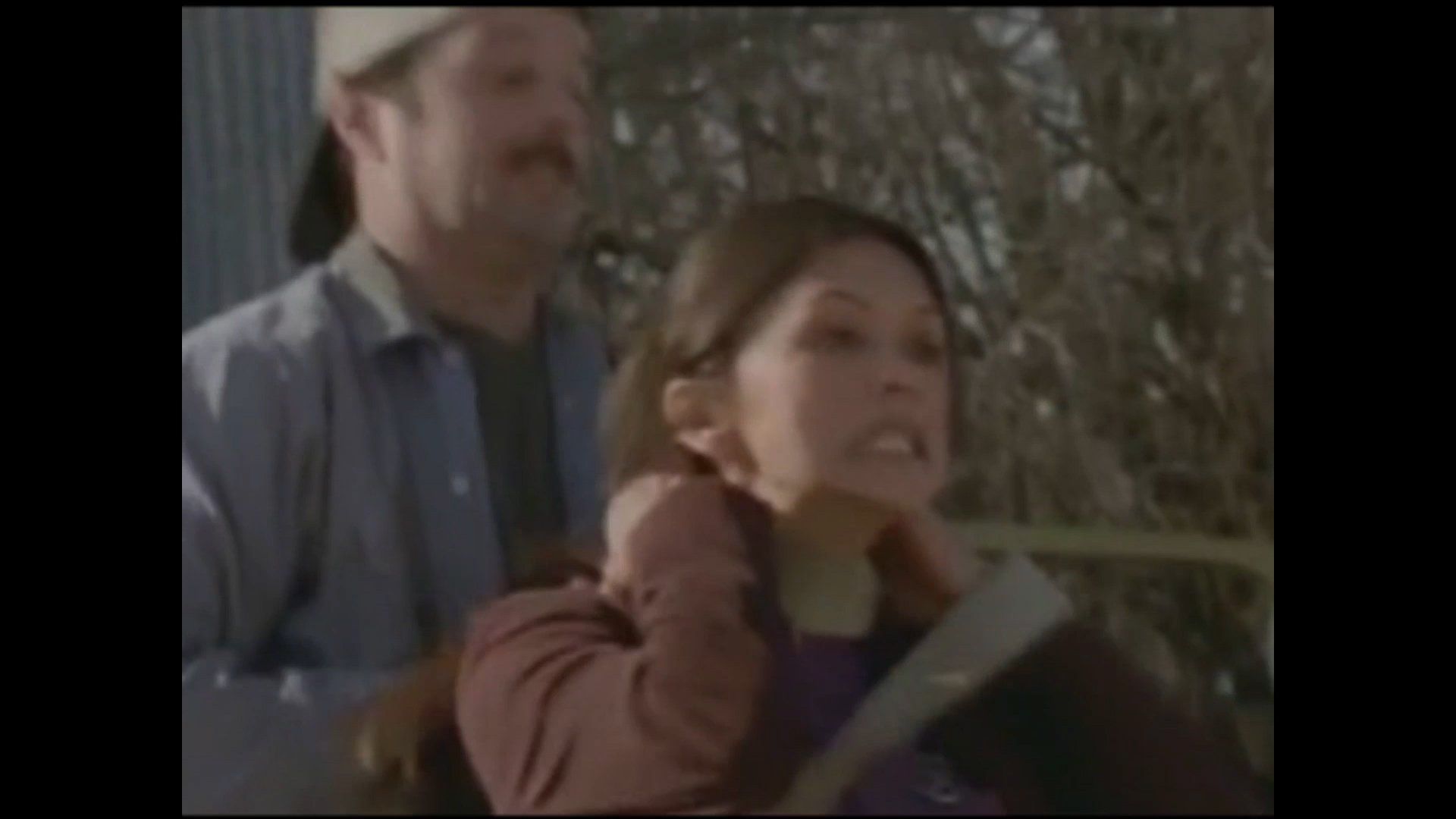 A person grips their own neck with a strained expression, while a person wearing a cap stands directly behind them.