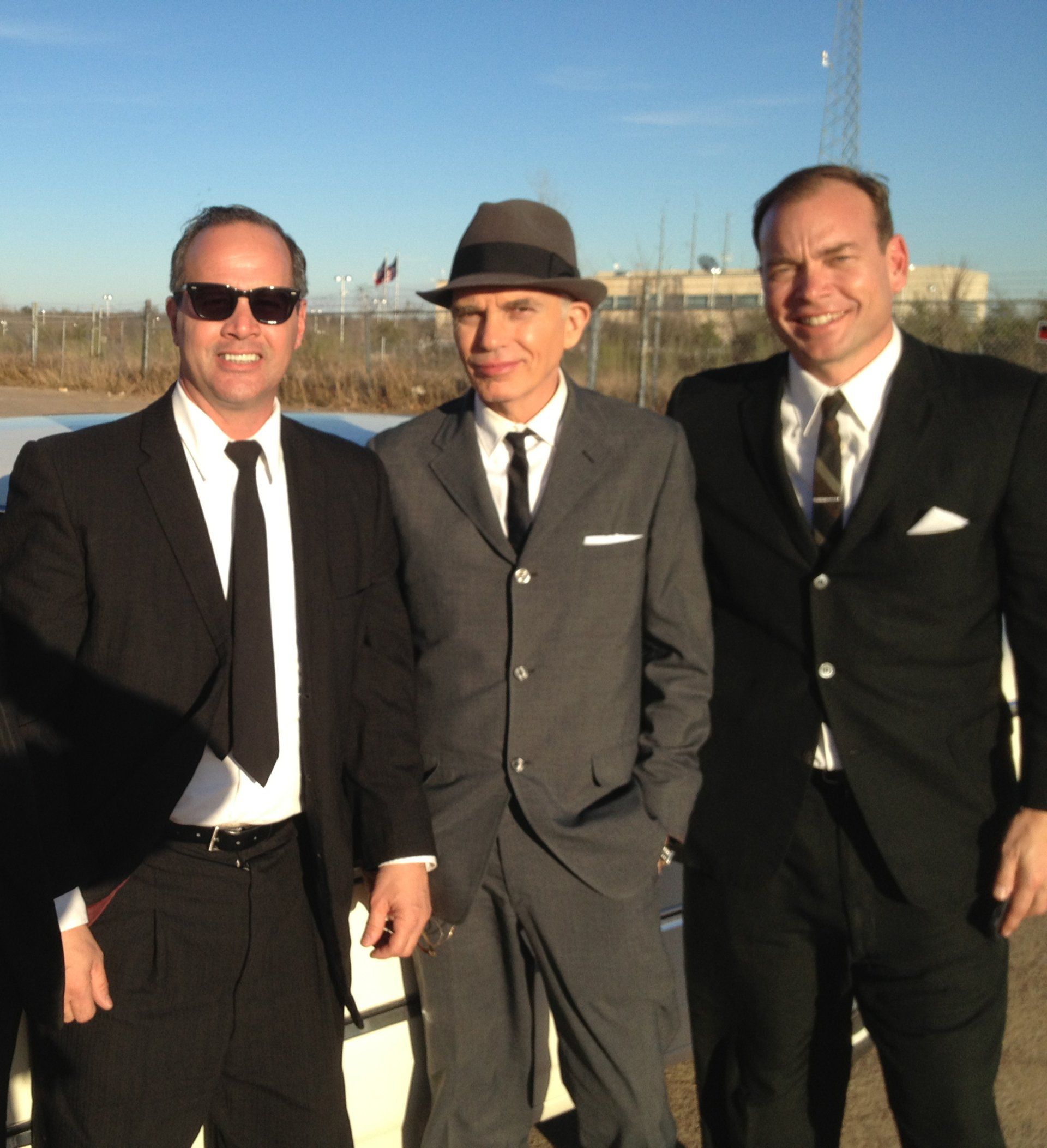 Three men in suits and ties pose for a photo outdoors, one wearing sunglasses and another wearing a fedora.