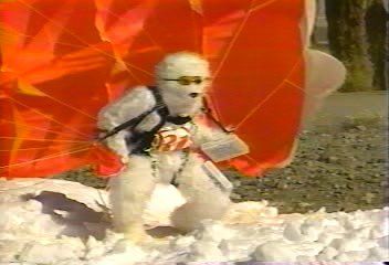 A white, bear-like figure in sunglasses and a harness stands in the snow beneath a large, orange parachute.