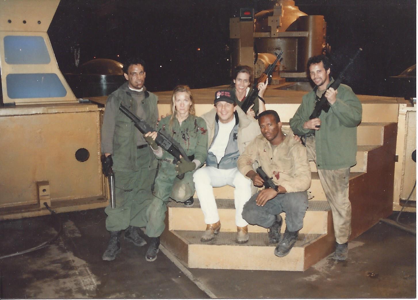 Six people posing on a film set with prop weapons, wearing military-style gear, including a central figure in a cap.