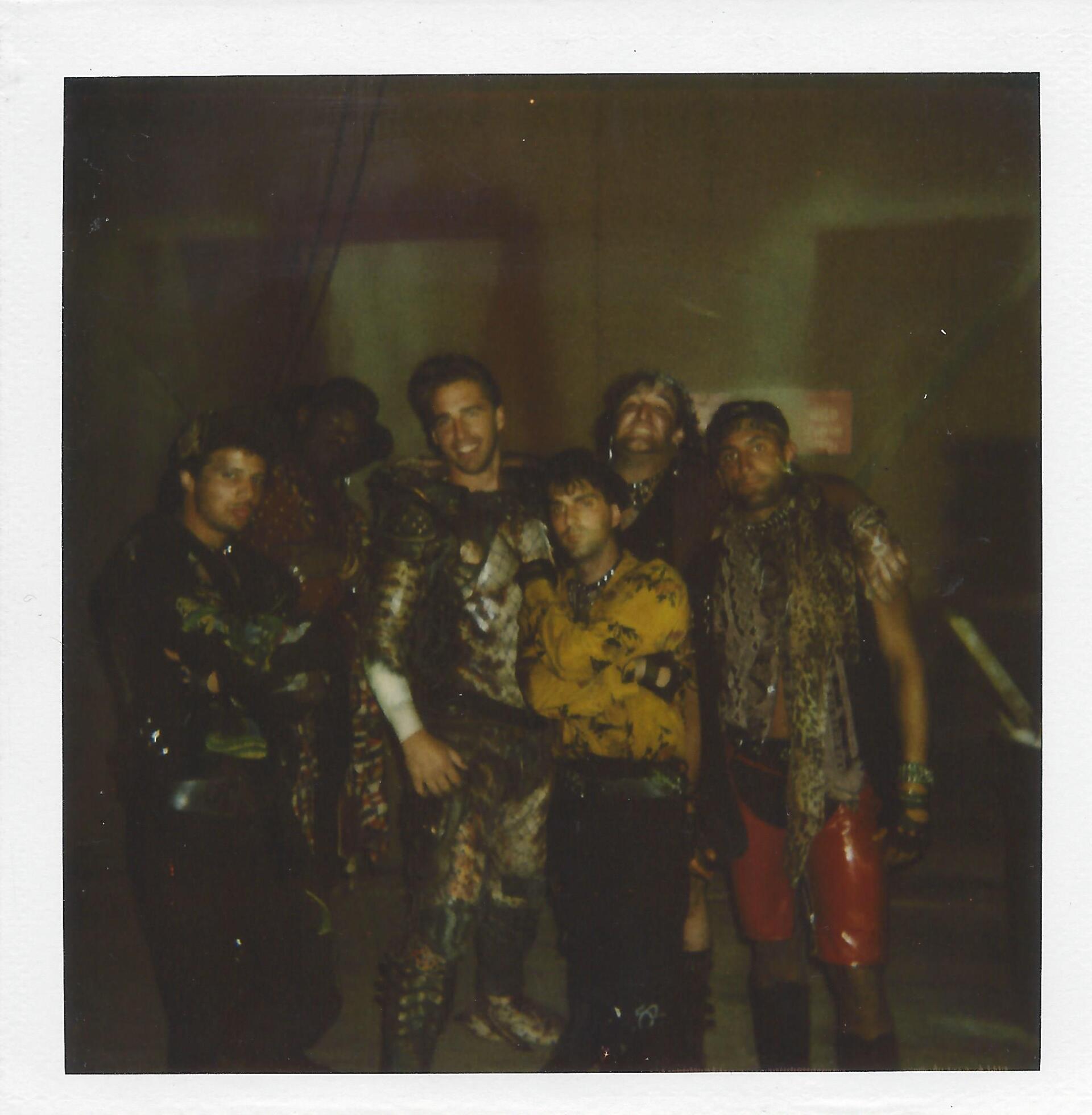 A group of six people in varied, dark-toned costumes pose together for a photograph in a dimly lit, indoor setting.