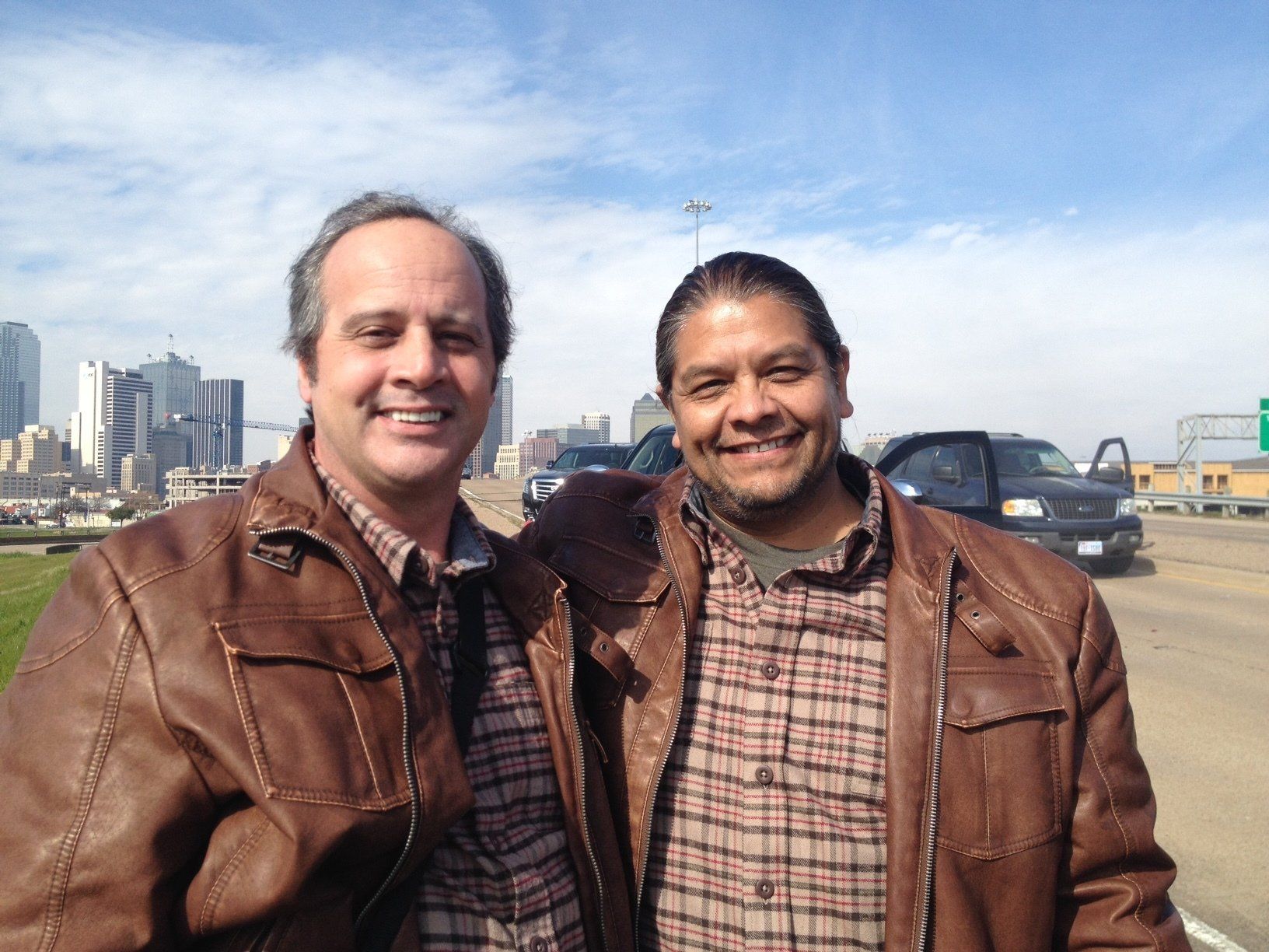 Two smiling men in brown leather jackets and plaid shirts stand side-by-side with a city skyline in the background.