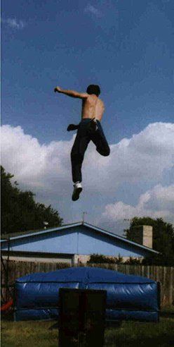 A person jumping high above a large blue inflatable landing mat outdoors against a bright blue sky.