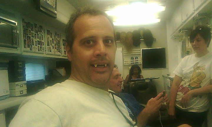 A man with prominent vampire-style teeth looks at the camera while others sit in a hair and makeup trailer.