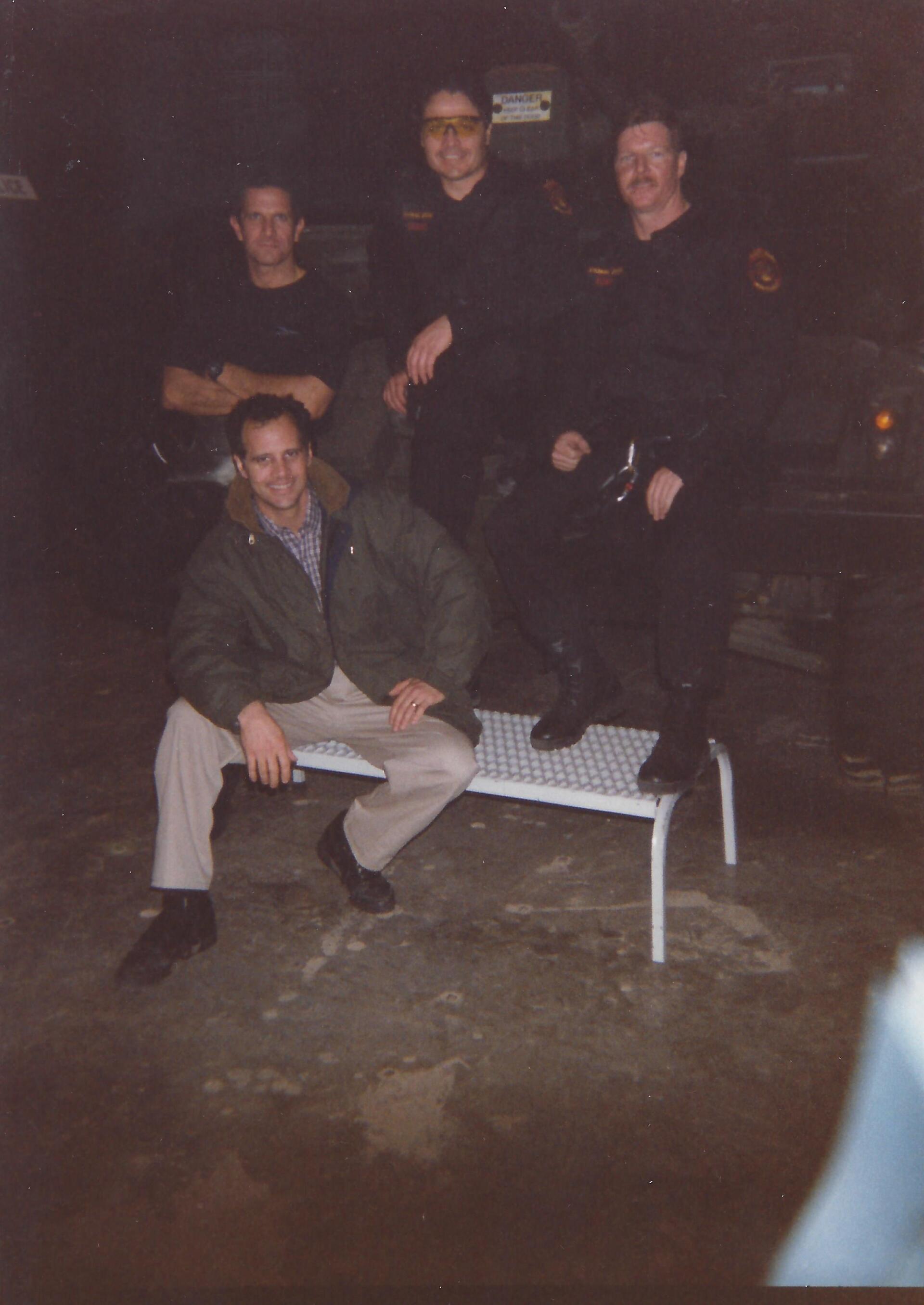 Four people pose for a group photo; one person sits on a low bench while the others stand or sit behind them indoors.