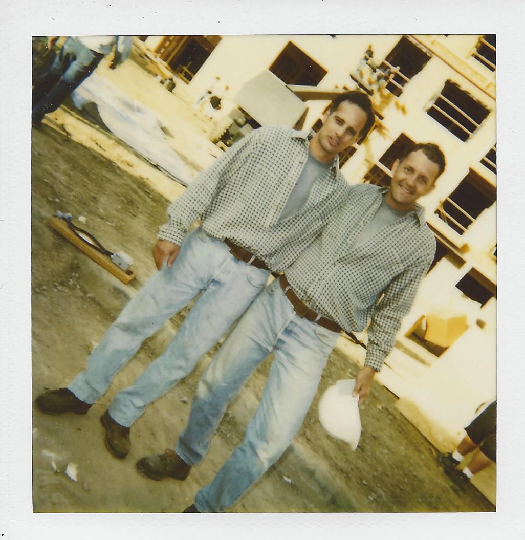 Two individuals in button-down shirts and jeans standing on a construction site, holding hard hats.