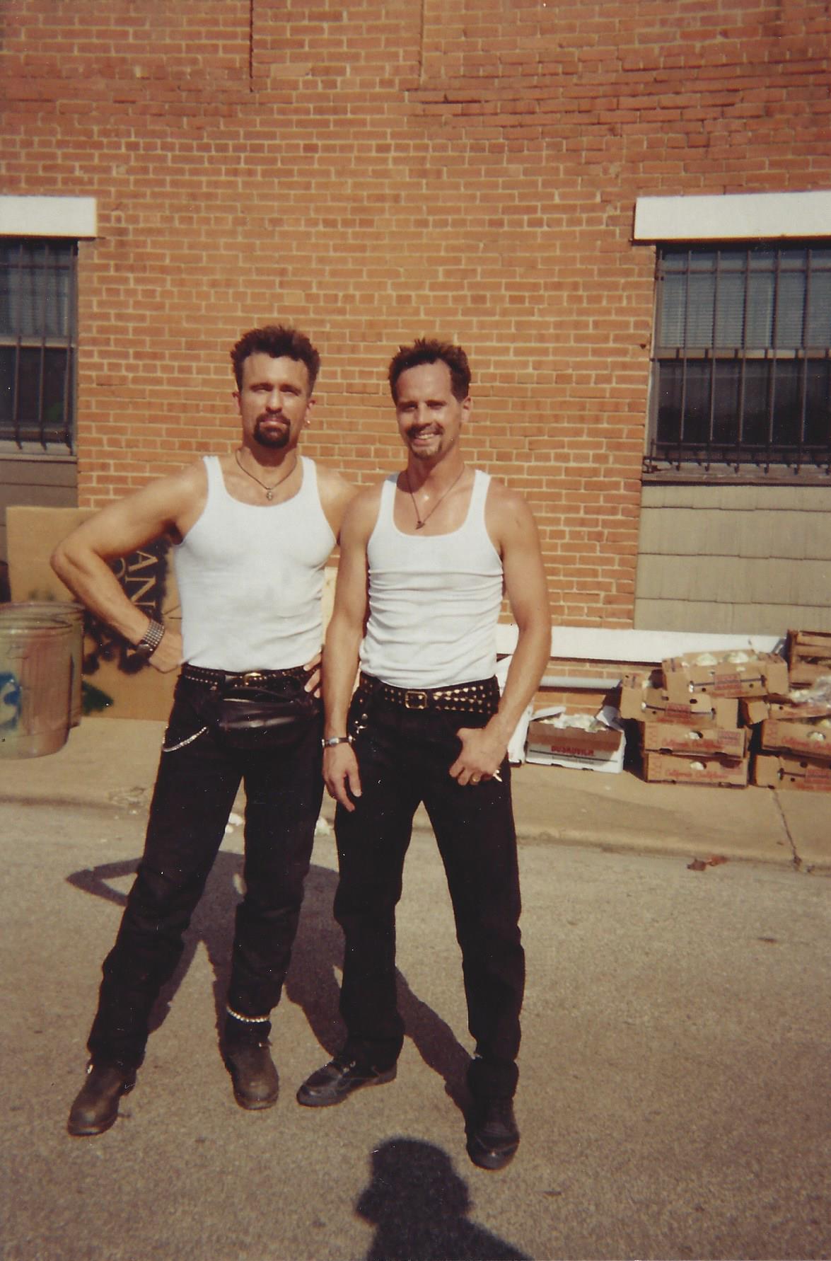 Two people wearing white tank tops and black pants stand side-by-side in front of a red brick building.