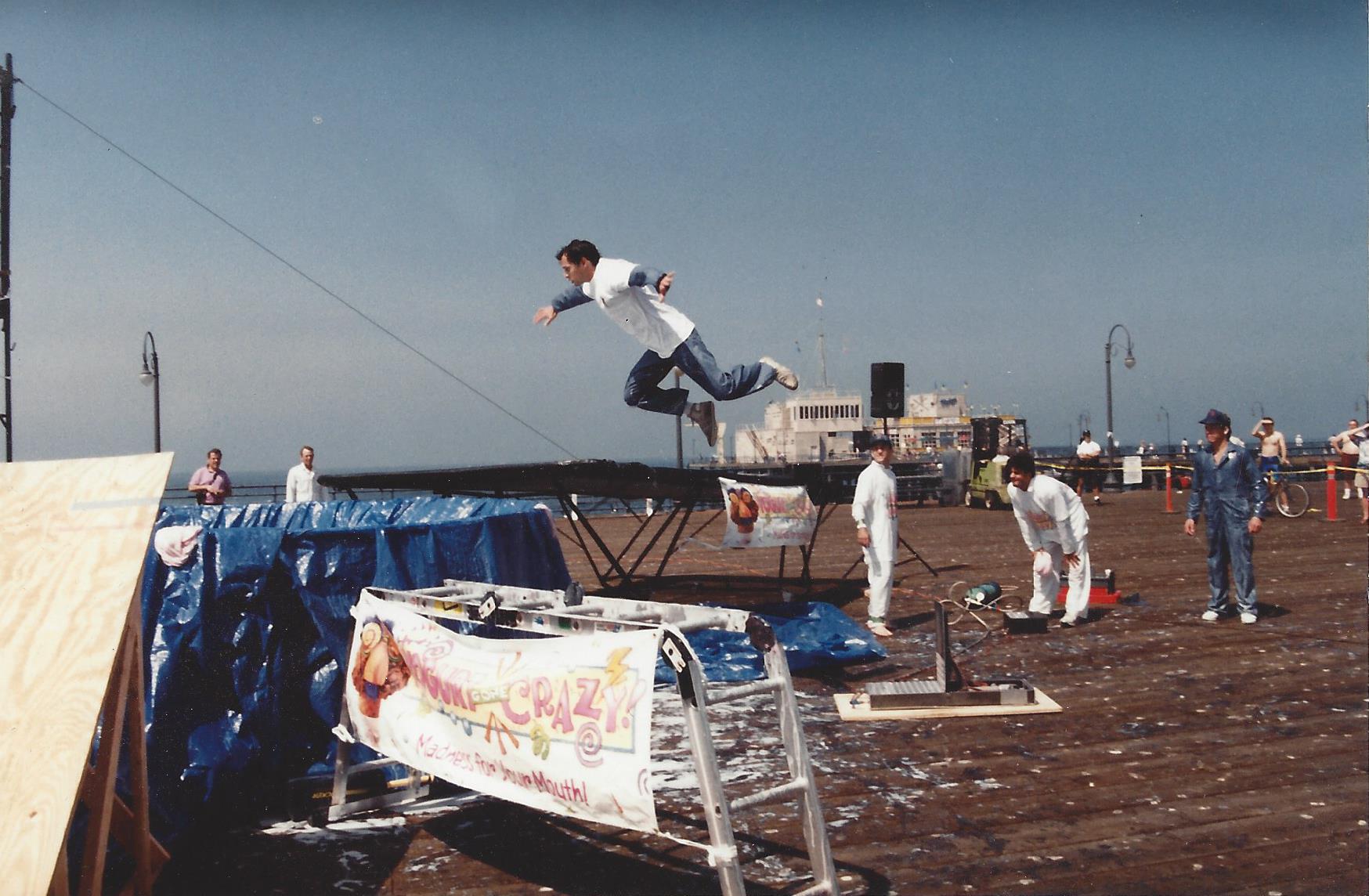 A person performs a mid-air jump from a trampoline on a wooden pier, with several spectators standing nearby.