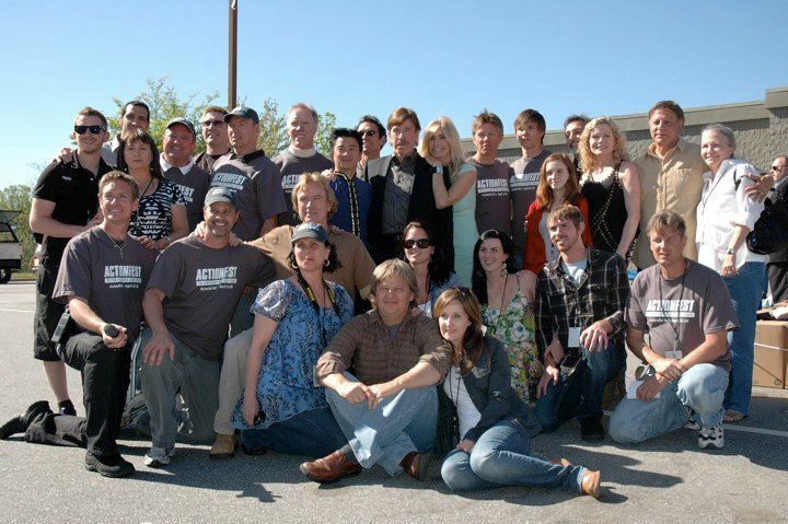 A large group of people poses for a sunny outdoor portrait, standing and sitting together in an asphalt parking lot.
