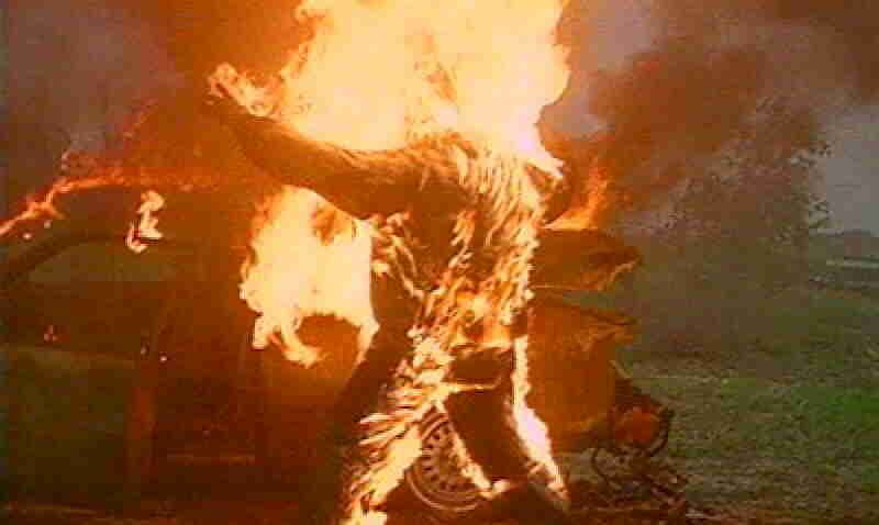 A person standing with arms outstretched while engulfed in bright orange flames in an outdoor, rural setting.