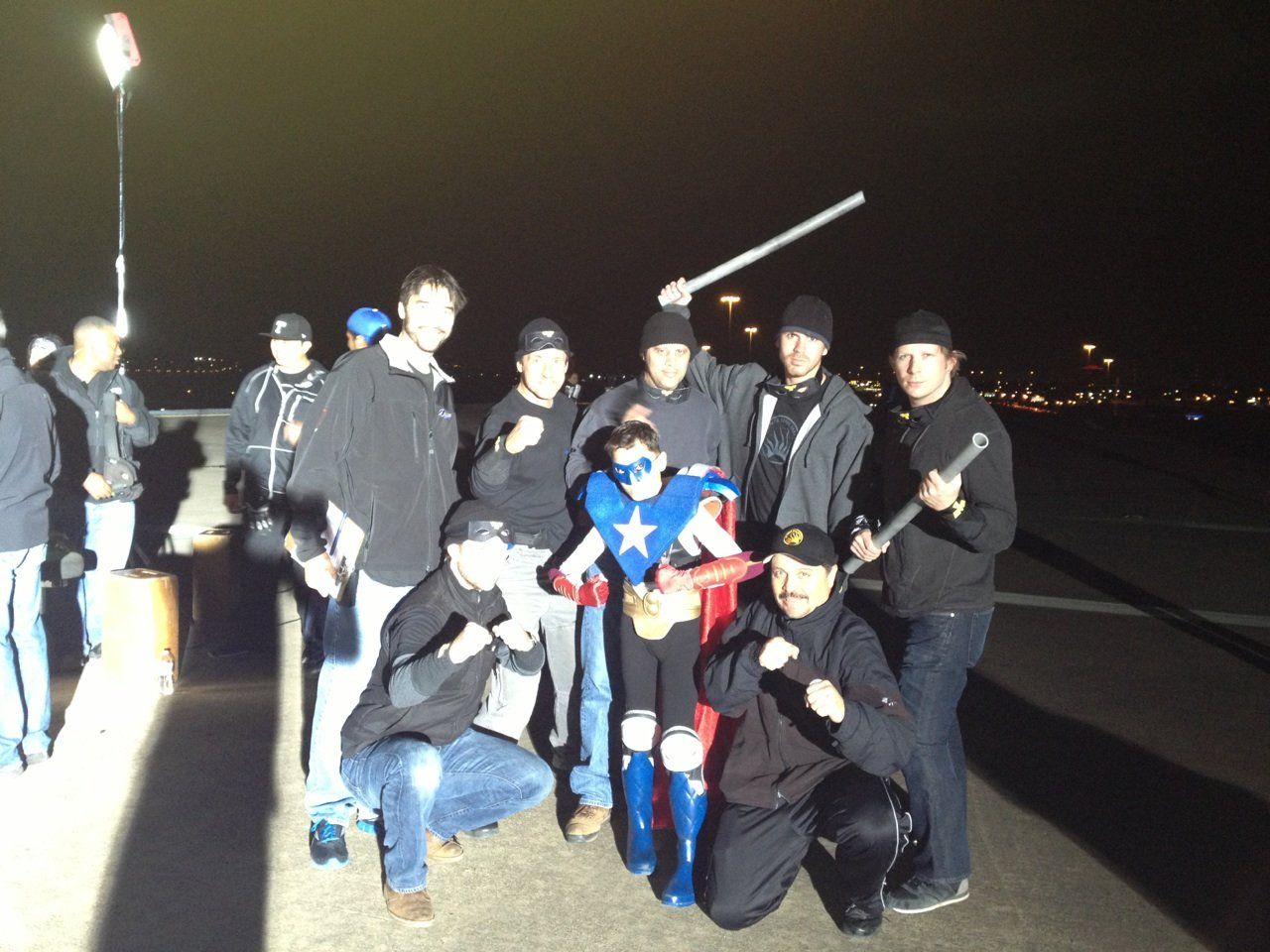 A group of people standing at night outdoors, some holding props, surrounding a person wearing a superhero costume.