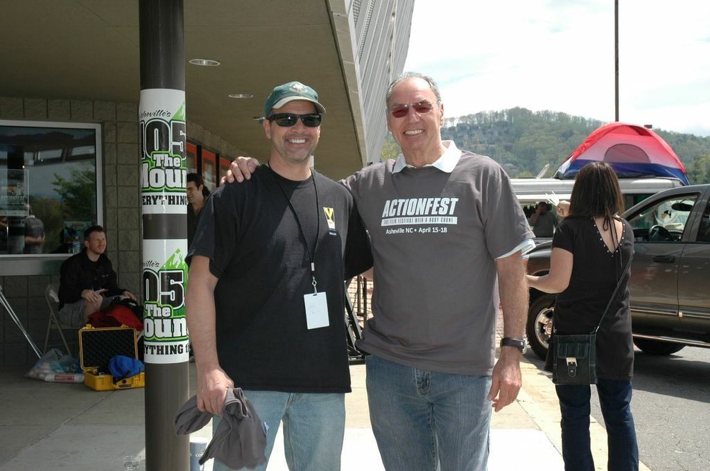 Two people stand smiling under a building overhang. One wears a black t-shirt and hat, the other a grey Actionfest shirt.