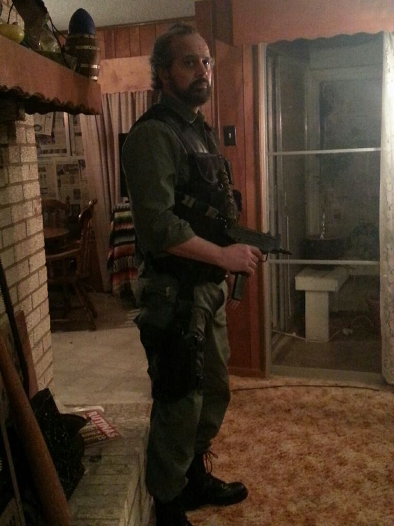 A person in a tactical vest and green uniform stands in a living room, holding a firearm.