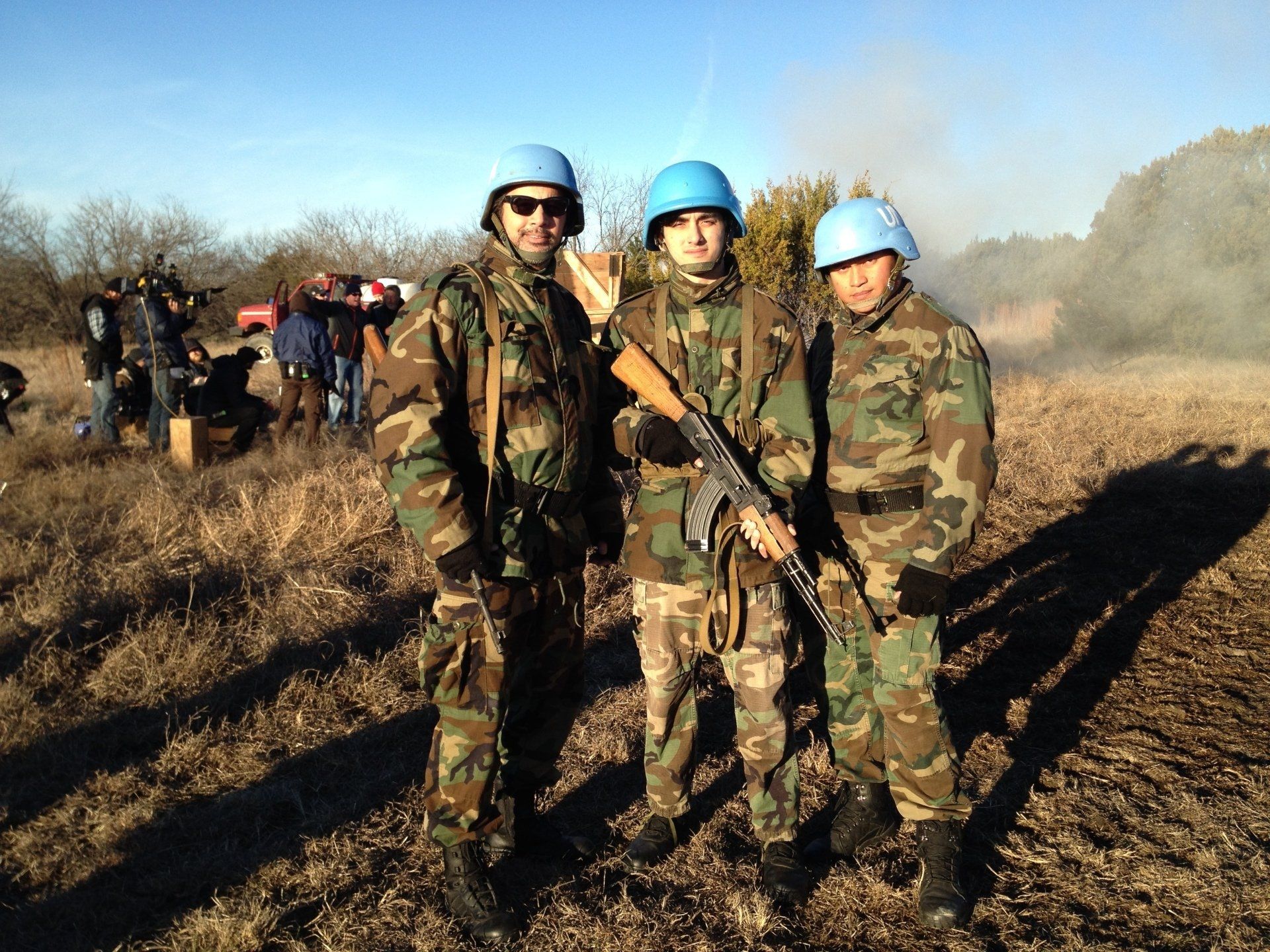 Three people in camouflage uniforms and blue helmets stand in a field, one holding a rifle, with others in the background.